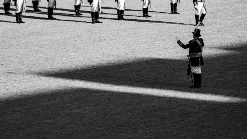 Officer directs soldiers in historical uniforms on a grassy field with strong shadows.