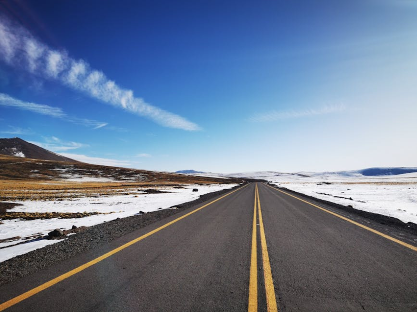Long road with yellow lines cuts through snowy, dry landscape under a wide blue sky.