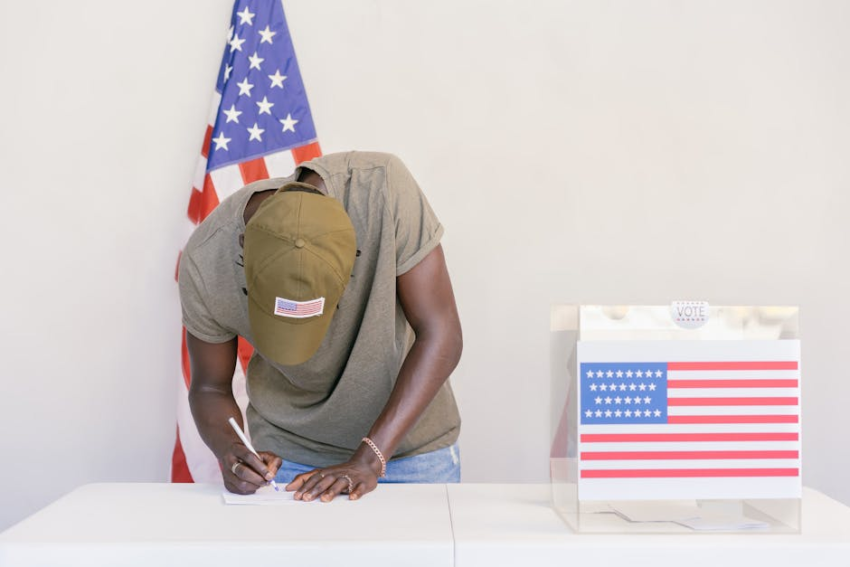 Black person in a hat writing on a document near an American flag and ballot box.