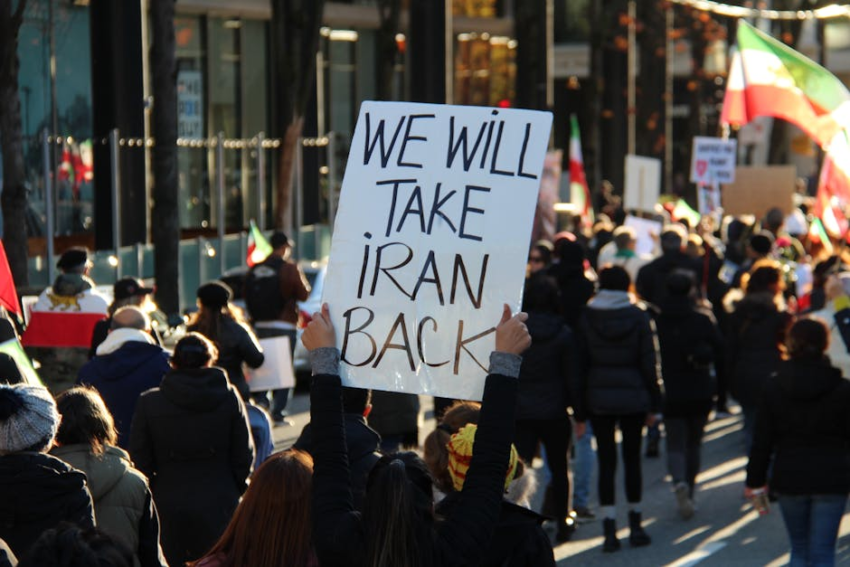 Protesters march with a sign reading 'WE WILL TAKE IRAN BACK' and pre-revolutionary Iranian flags.