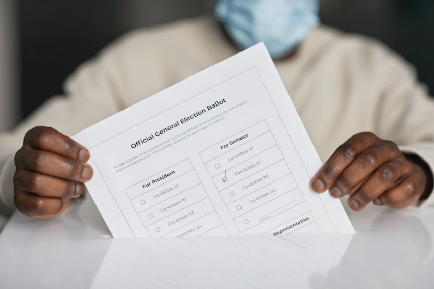 Person with dark skin wearing a mask places a marked election ballot into a ballot box.