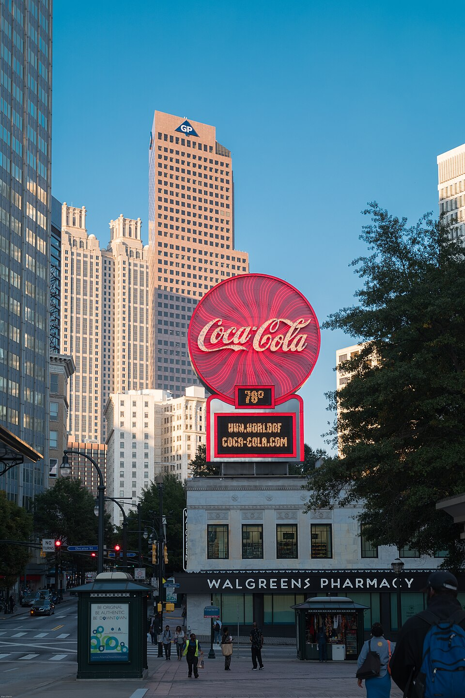 Street-level photo: View of the landmark Coca-Cola neon sign atop the recently restored Olympia Building and its new Walgreens, taken from near the Five Points MARTA Station. The 17-story Beaux-Arts Candler Building, 52-story pink granite Georgia Pacific Tower, and the 50-story postmodern 191 Peachtree Tower can be seen on the far side of Woodruff Park in the background.