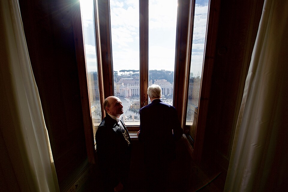 Street-level photo: U.S. Secretary of State John Kerry looks out a window at the Christmas tree in the middle of St. Peter's Square on December 2, 2016, before a one-on-one meeting with Pope Francis in the Papal Apartments at the Vatican in Vatican City. [State Department photo/ Public Domain]
